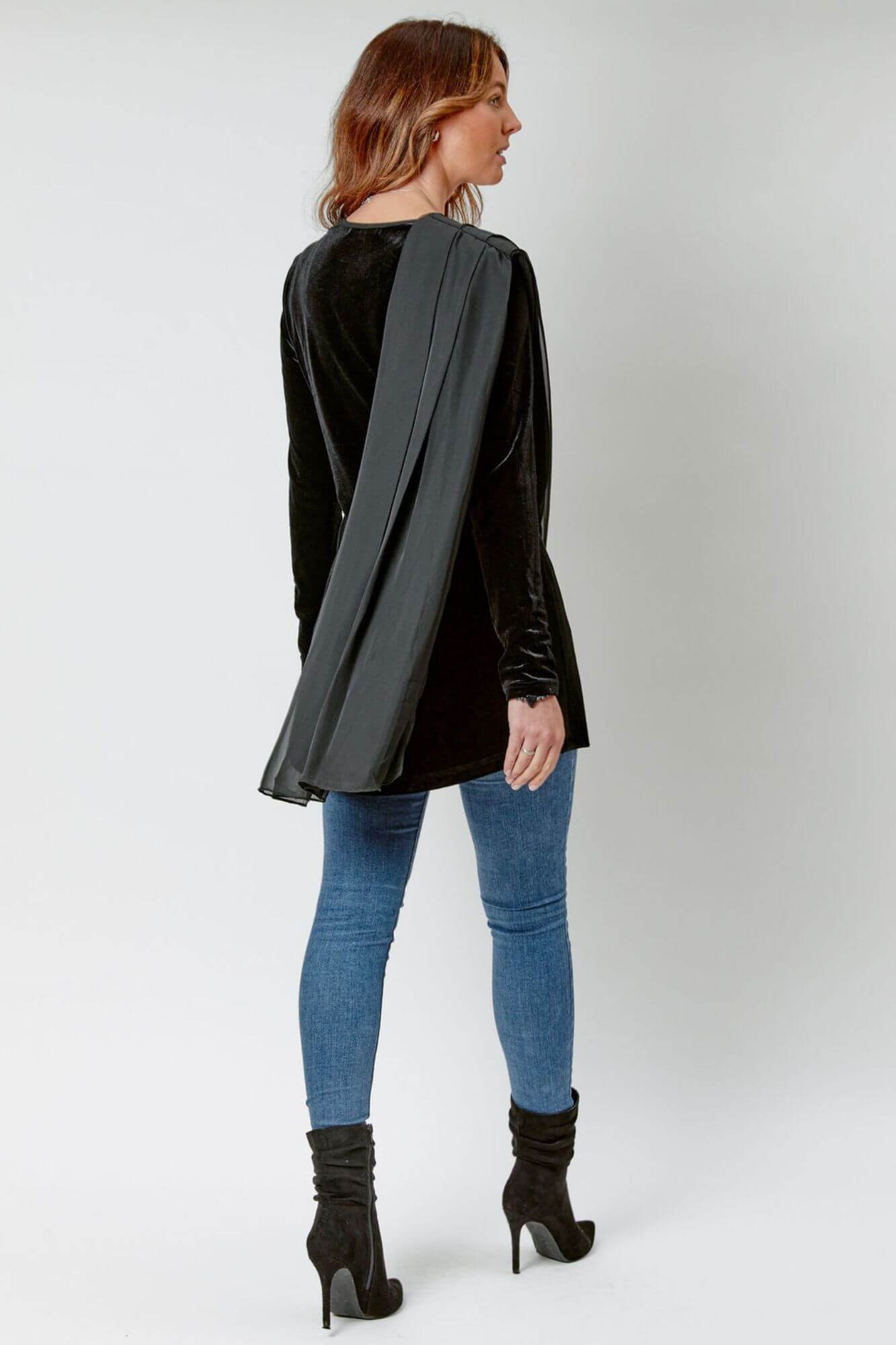 Woman wearing a black long-sleeve top with a wide shoulder strap, blue jeans, and black high-heeled boots on a white background.