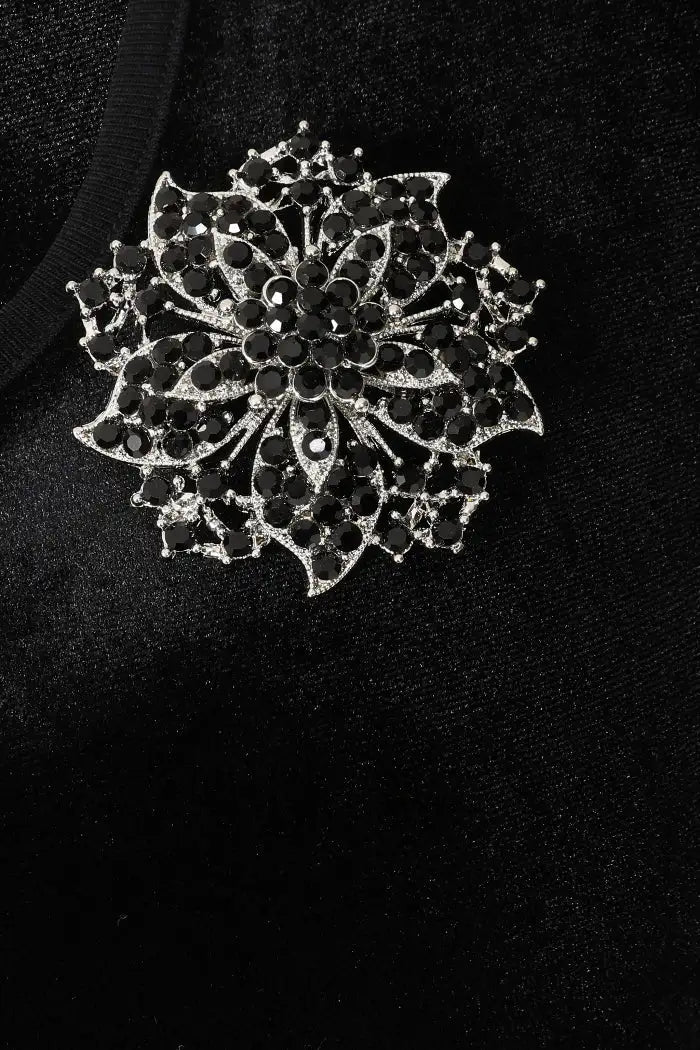 A silver-toned floral brooch embellished with black rhinestones, displayed on a textured black fabric background.